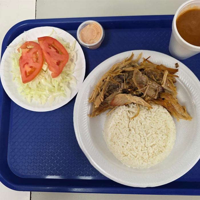 Bandeja azul con plato de pernil deshebrado y arroz blanco, ensalada de repollo con tomate y vaso de habichuelas